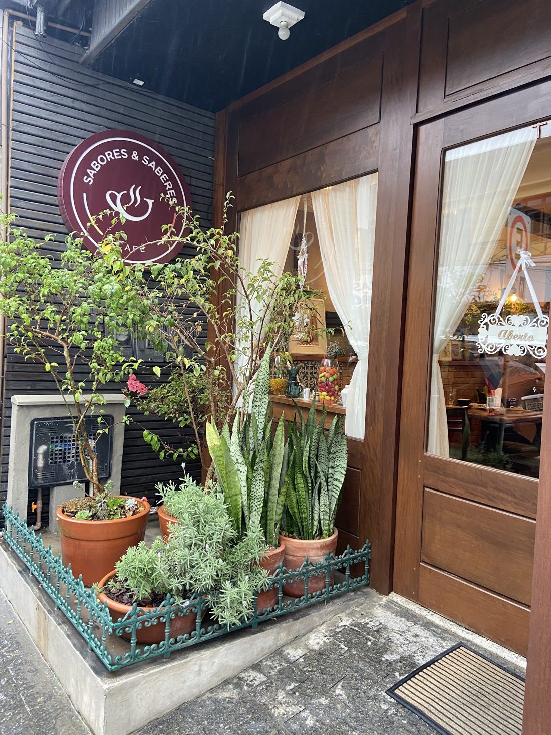 Entrada do café com a placa redonda, vasos de plantas e cortinas na janela