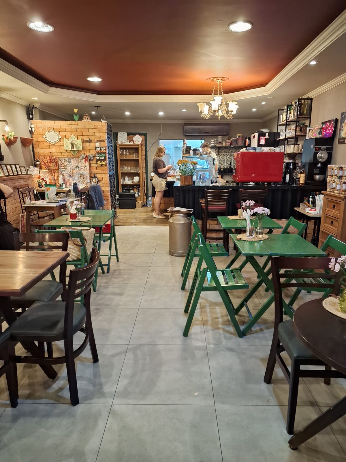 Salão do café com cadeiras verdes, parede de tijolinhos e balcão ao fundo